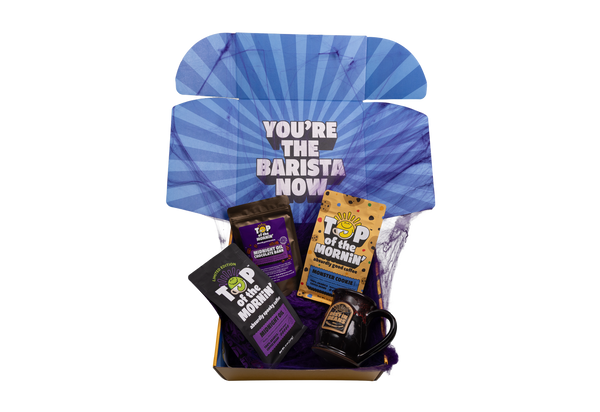 A curated coffee gift box set against a white background, featuring a blue and purple sunburst-patterned inner lid with the text 'YOU'RE THE BARISTA NOW', along with various coffee accessories including 'Top of the Morning' branded mugs and bags.